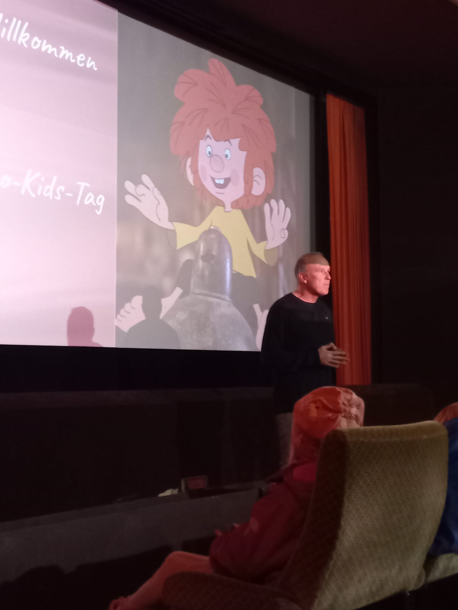Felix Zosel begrüßt die Kinder und ihre Eltern auf der Bühne. Im Hintergrund ist Pumuckl auf der Leinwand zu sehen.